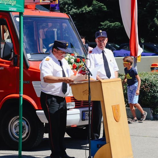 Zwei Männer in Uniformen stehen an einem Rednerpult vor einem Feuerwehrauto. Einer liest eine Rede, während der andere zuhört. Ein Junge beobachtet von der Seite.