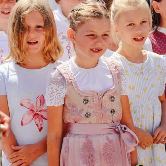 Drei junge Mädchen, wahrscheinlich in einer Schulumgebung, stehen zusammen. Das mittlere Mädchen in einem Blumenkleid lächelt in die Kamera, während die anderen Freizeitkleidung tragen.