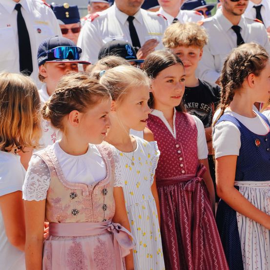 Eine Gruppe junger Mädchen in traditionellen Kleidern steht zusammen. Einige haben Zöpfe, andere kurze Haare. Sie sind wahrscheinlich bei einer Schulveranstaltung. Im Hintergrund beobachten Menschen in Uniformen und Mützen.