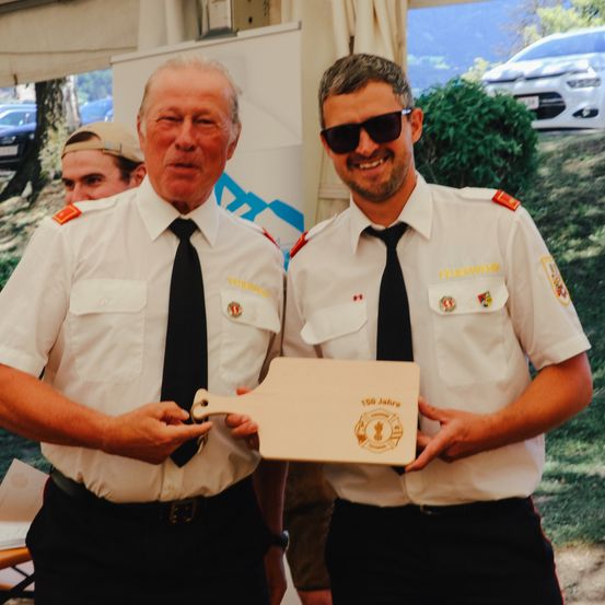 Zwei Männer in weißen Uniformen halten eine Plakette mit der Aufschrift 150 Jahre und stehen nebeneinander mit lächelnden Gesichtern.