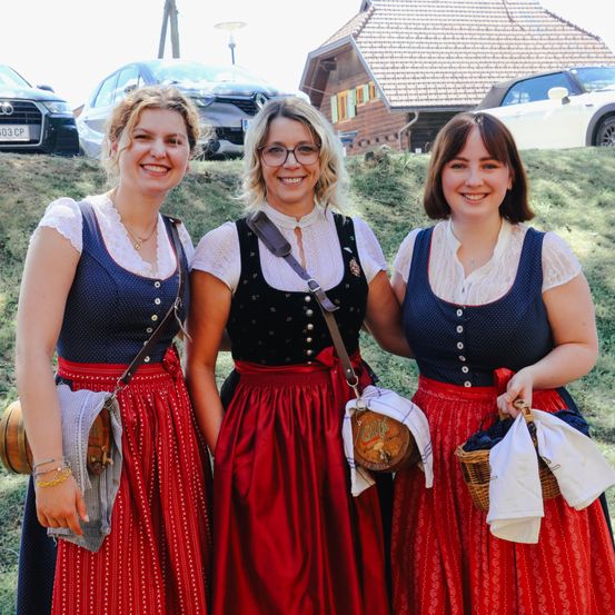 Drei Frauen in traditioneller Kleidung stehen lächelnd zusammen, mit Körben und Taschen. Ein Haus und Autos sind im Hintergrund.