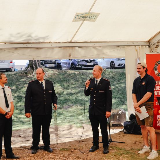 Vier Männer in Uniformen stehen unter einem Zelt. Einer hält ein Mikrofon. Fahrzeuge sind auf einem Grashügel hinter ihnen geparkt. Ein Banner ist rechts.