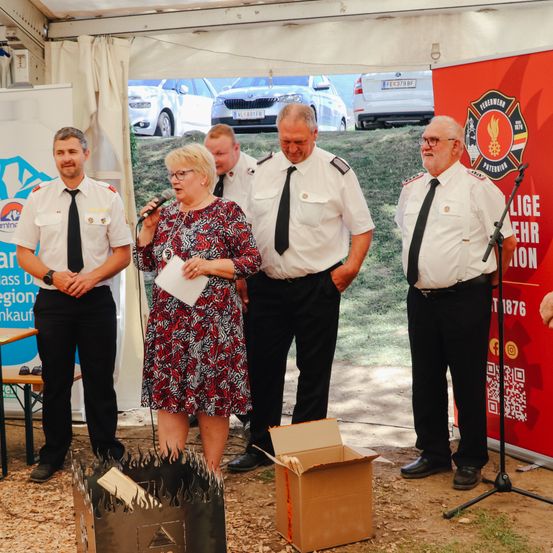 Eine Frau spricht in ein Mikrofon bei einer Veranstaltung. Drei Feuerwehrleute stehen hinter ihr, und ein Feuerlöscher ist in der Nähe. Ein roter Banner und Autos sind im Hintergrund zu sehen.