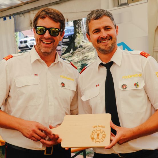 Zwei Feuerwehrleute halten eine Holzplakette mit der Aufschrift 150 Jahre, stehen unter einem Zelt im Freien.