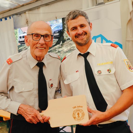 Zwei Feuerwehrleute in Uniform halten eine Plakette. Ein Mann trägt eine Brille und einen Abzeichen. Der andere lächelt. Hinter ihnen steht ein Zelt mit weißem Hintergrund.