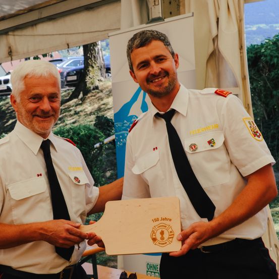 Zwei Feuerwehrleute, einer mit einer Plakette, lächeln und schauen zur Kamera. Ein Mann hält eine Plakette mit Logo und Text, 150 Jahre.
