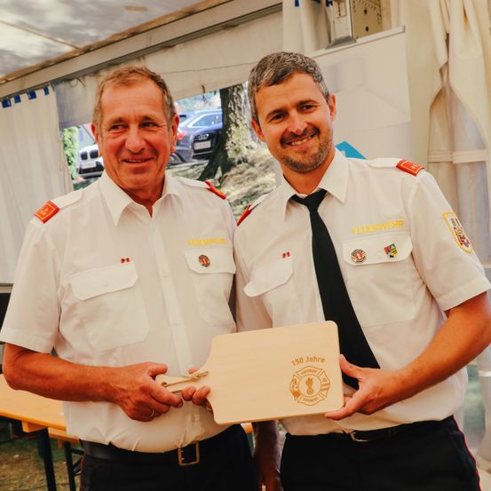 Zwei Männer in Uniform stehen und halten eine Plakette mit der Aufschrift 150 Jahre. Sie scheinen Feuerwehrleute zu sein.