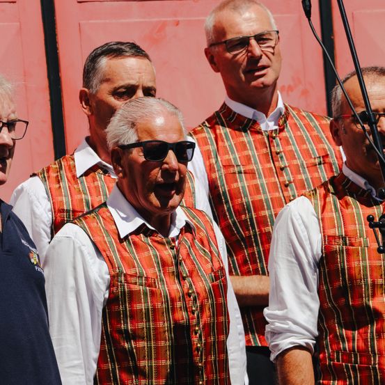 Eine Gruppe von Männern in karierten Westen, einige mit Sonnenbrille, stehen vor einer roten Tür. Ein Mann in der Mitte lacht. Ein Mikrofon auf einem Ständer ist im Vordergrund zu sehen.