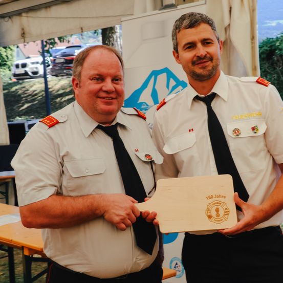 Zwei Feuerwehrleute halten eine Holzplakette mit der Aufschrift 150 Jahre. Sie stehen lächelnd unter einem Zelt.