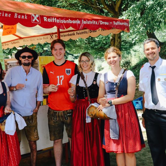 Eine Gruppe von Menschen, darunter ein Mann in einem roten Shirt, steht vor einem Zelt mit einem Schild. Frauen in traditioneller Kleidung halten Getränke.