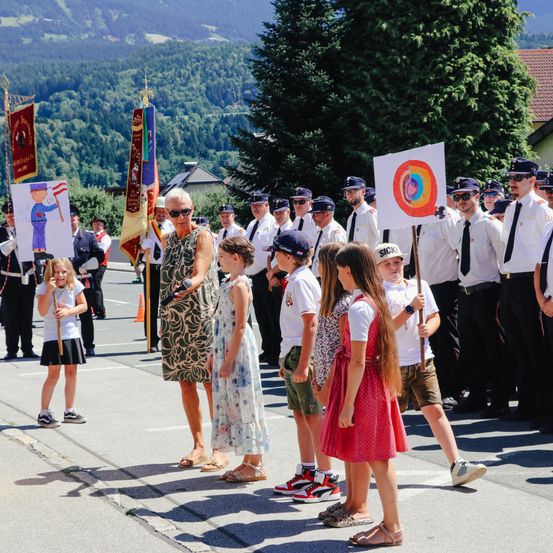 Eine Gruppe von Kindern und Erwachsenen steht in einer Reihe auf der Straße mit Bergen im Hintergrund und hält Fahnen und Schilder.