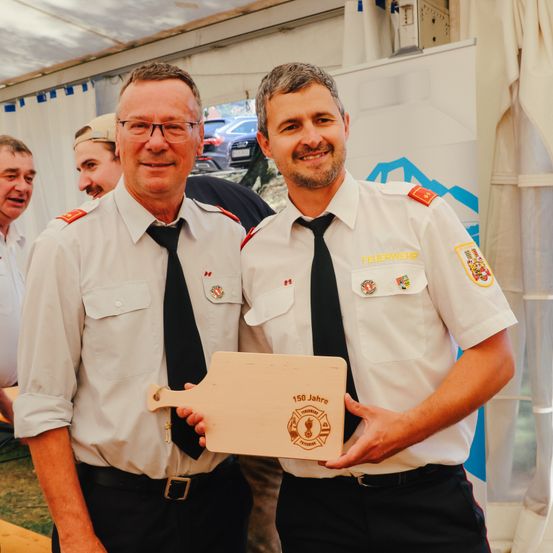 Zwei Männer in Feuerwehruniformen halten eine Holzplakette. Sie lächeln und scheinen für ein Foto zu posieren.