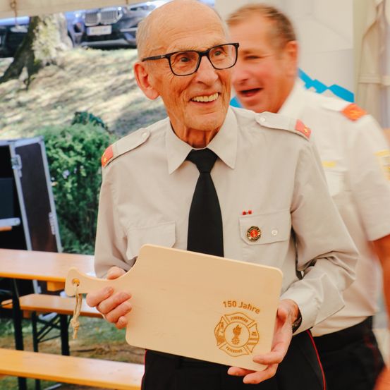 Ein älterer Mann in Uniform hält eine Plakette, die 150 Jahre feiert. Ein anderer Mann in Uniform steht lächelnd hinter ihm. Sie sind bei einer Outdoor-Veranstaltung, mit Tischen und Bänken im Hintergrund.