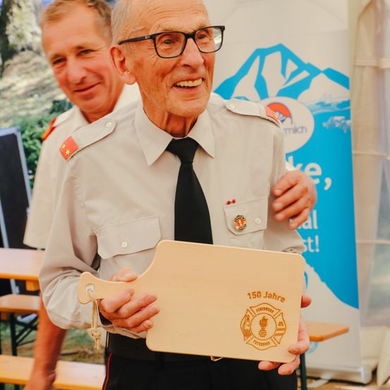 Ein älterer Mann in Uniform und mit einem Holzschild mit Text lächelt und steht neben einem anderen Mann.