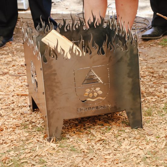 Ein Metall-Feuerkorb mit Flammendesign und Holzverbrennung, umgeben von Asche auf dem Boden, mit einem Logo vorne.