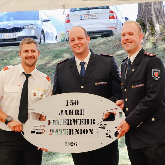 Drei Feuerwehrleute halten eine Plakette für das 150-jährige Jubiläum der Feuerwehr Paternion, mit einem Auto und einem Baum im Hintergrund.