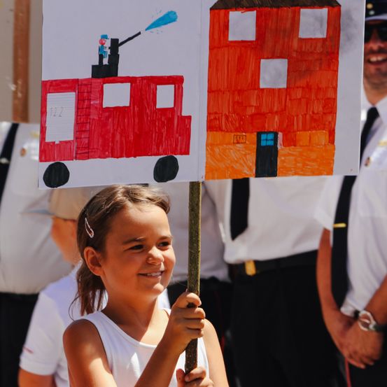 Ein Mädchen hält ein Schild mit einer Feuerwehrwagen- und Gebäudezeichnung und lächelt, während sie zwischen uniformierten Männern steht.