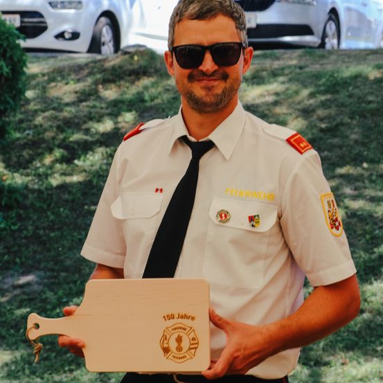 Ein Feuerwehrmann in Uniform hält eine Holzplakette, die 150 Jahre Dienst feiert. Er lächelt für die Kamera, mit einem Auto und Grünpflanzen im Hintergrund.