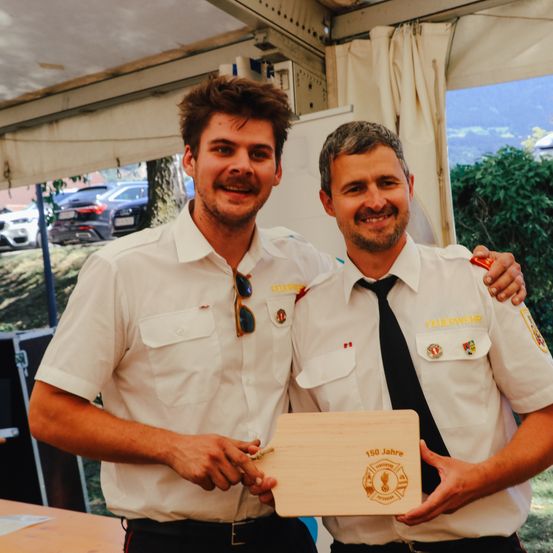 Zwei Feuerwehrleute in Uniform, einer hält eine Plakette mit Logo und '150 Jahre' Text, stehen lächelnd zusammen draußen, mit Autos und Bergen im Hintergrund.