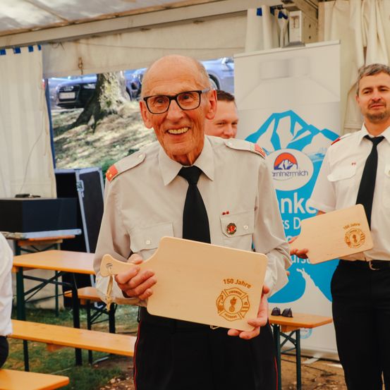 Ein älterer Mann in Uniform hält eine Holztafel mit der Aufschrift 150 Jahre. Er lächelt und steht vor einem weißen Banner mit Logo. Ein weiterer Mann in Uniform steht hinter ihm und hält eine ähnliche Tafel.