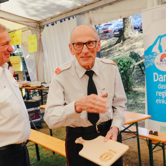 Ein älterer Mann in Uniform hält ein Holzschneidebrett und steht vor einem Zelt mit einem anderen Mann in der Nähe. Dahinter ist ein Tisch mit verschiedenen Gegenständen zu sehen.