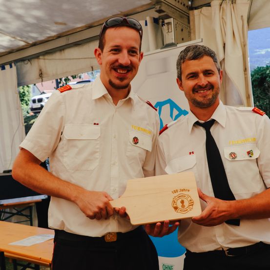 Zwei Feuerwehrleute in Uniform halten eine Plakette zur Feier von 150 Jahren Dienst. Sie lächeln und posieren unter einem Zelt.