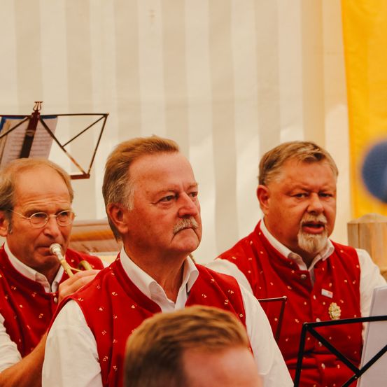 Drei Männer in roten Westen spielen, einer hält eine Trompete. Der Mann in der Mitte sieht ernst aus, während die anderen auf ihre Instrumente konzentriert sind.