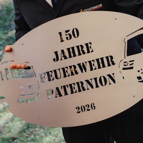 Eine Person hält eine ovale Plakette in Form eines Feuerwehrwagens, die das 150-jährige Jubiläum der Paternon Feuerwehr feiert, datiert auf 2026.