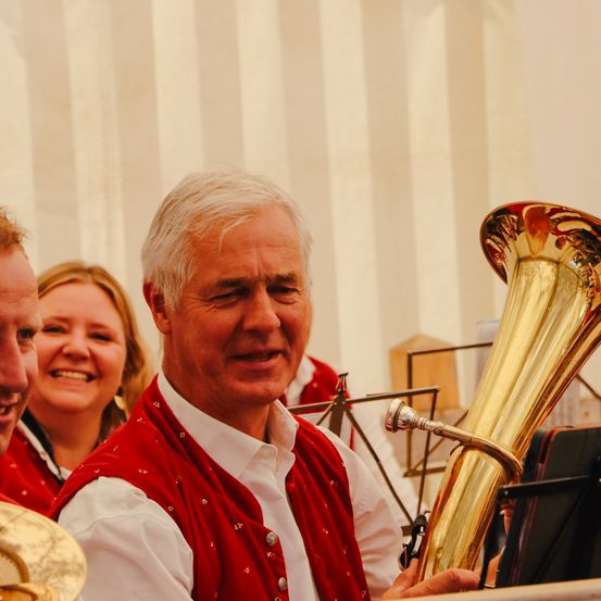 Ein Mann in einem roten Weste lächelt, während er eine große Tuba hält. Eine Frau lächelt hinter ihm, und ein anderer Mann schaut auf der linken Seite. Sie scheinen Teil einer Band zu sein.