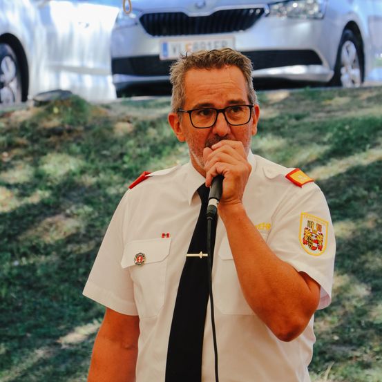 Ein Mann in einer weißen Uniform spricht mit ernster Miene in ein Mikrofon. Ein Auto ist im Hintergrund verschwommen.