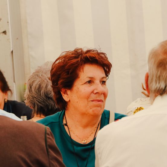 Eine Frau mit roten Haaren und einem grünen Shirt lächelt bei einem Treffen. Ein älterer Mann in einem weißen Shirt ist in der Nähe.
