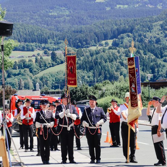 Eine Marschkapelle steht auf einer Straße, hält Fahnen und hat einen roten Feuerwehrwagen und Berge im Hintergrund.