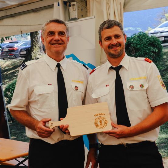 Zwei Feuerwehrleute, beide in Uniform, halten eine Holzplakette mit der Aufschrift 150 Jahre. Sie lächeln und stehen draußen.