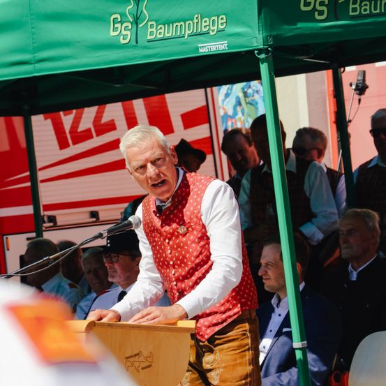 Ein Mann steht an einem Rednerpult und spricht in ein Mikrofon unter einem grünen Zelt, trägt eine rote Weste. Viele Leute sind im Publikum, einige tragen Sonnenbrillen und Westen. Ein großes Banner hinter dem Mann ist teilweise sichtbar.