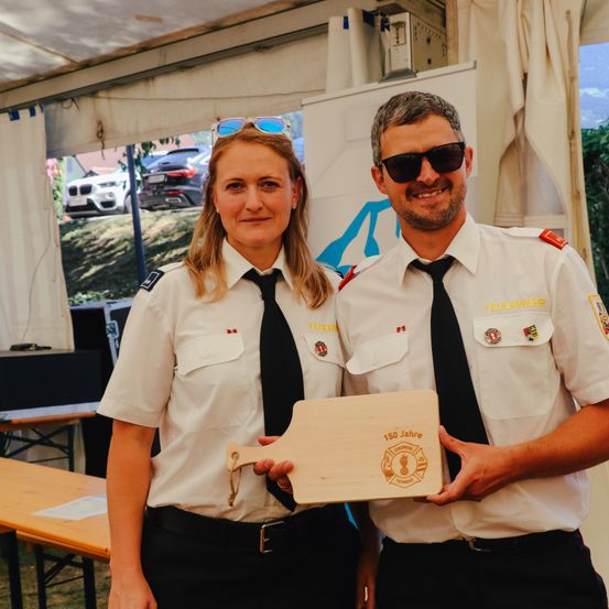 Zwei Personen in Uniform stehen unter einem Zelt und halten eine Holztafel mit der Aufschrift '150 Jahre.' Sie lächeln, und im Hintergrund sind Autos und Grünfläche zu sehen.