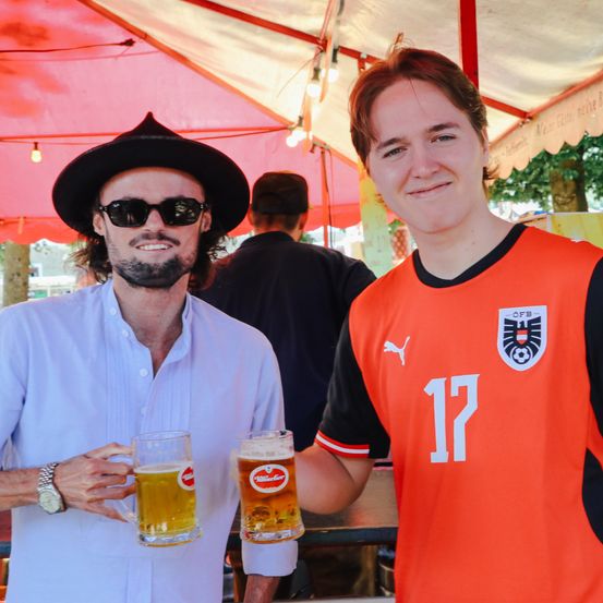 Zwei Männer halten Biergläser unter einem Zelt mit rotem Dach, einer trägt ein blaues Hemd und einen Hut, der andere ein oranges Trikot mit der Nummer 17.