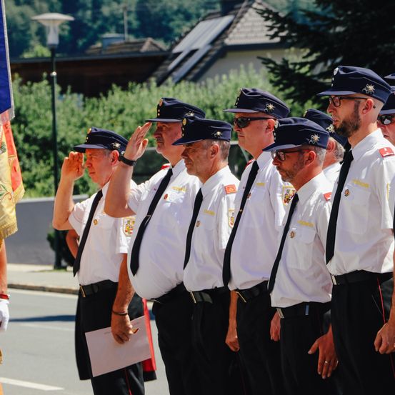 Uniformierte Männer, wahrscheinlich bei einer feierlichen Veranstaltung, stehen in einer Reihe und salutieren. Die Männer tragen Hüte, Krawatten und Abzeichen. Im Hintergrund befinden sich Bäume, ein Haus und eine Straßenlaterne.