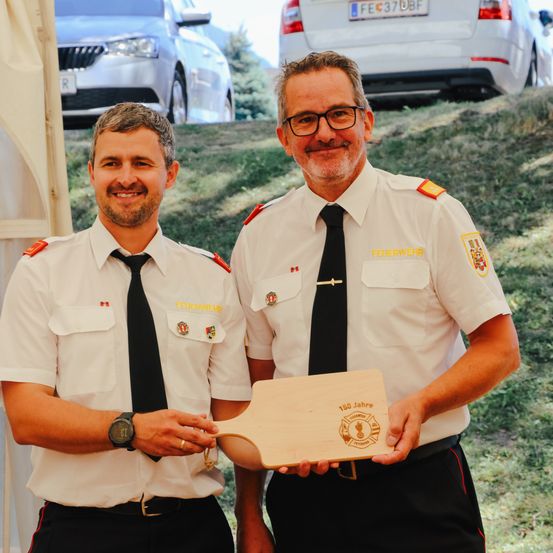 Zwei Männer in Uniformen stehen und lächeln, einer hält eine Holzplakette. Der Mann auf der linken Seite trägt eine Uhr und einen Ring. Hinter ihnen stehen Autos auf einem Hang geparkt.