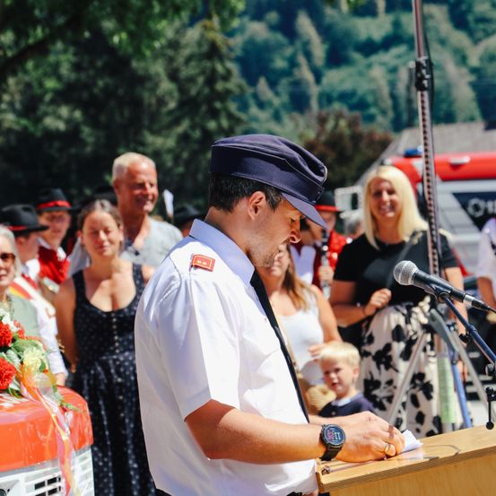 Ein Mann in Uniform spricht an einem Podium vor einem Publikum. Dahinter schauen Menschen zu, darunter eine Frau mit blonden Haaren und einem Kind.