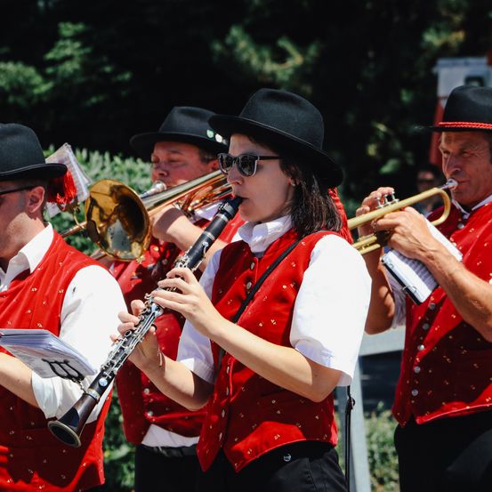 Eine Gruppe von Musikern in roten Westen und schwarzen Hüten spielt im Freien Instrumente wie Trompeten und Posaunen in einem Park.