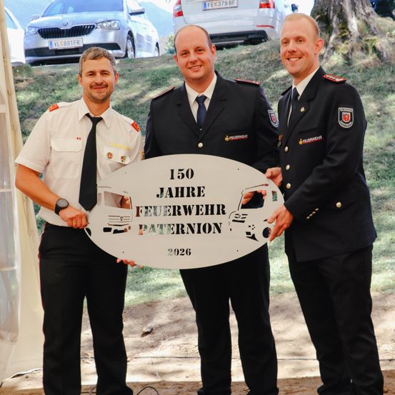 Drei Feuerwehrleute in Uniform stehen und posieren für ein Foto mit einem großen Schild, auf dem 150 Jahre Feuerwehr Paternion 2026 steht.