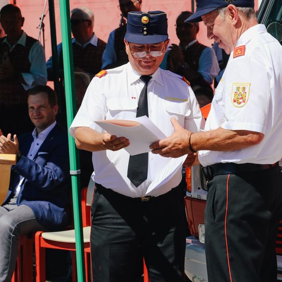 Zwei Männer in Uniform unterhalten sich vor einem Fahrzeug, einer hält ein Papier in der Hand, eine Menge beobachtet.