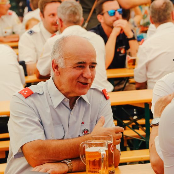 Ein älterer Mann in einer Uniform mit einem roten Stern auf seiner linken Schulter sitzt in einem Bierzelt, lächelt und schaut nach links. Er hat zwei Gläser Bier auf dem Tisch vor sich. Hinter ihm sitzen andere Menschen in Uniformen an Holztischen.