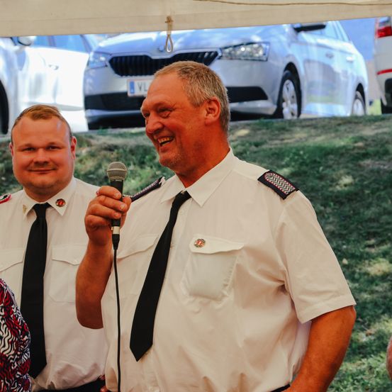 Zwei Männer in Uniformen stehen in einem Außenbereich. Der Mann vorne lächelt und hält ein Mikrofon, und er hält möglicherweise eine Rede. Hinter ihm steht ein weiterer Mann, der ebenfalls lächelt. Ein Auto ist hinter ihnen geparkt.