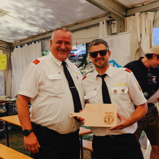 Zwei Feuerwehrleute in Uniform halten eine Plakette mit einem Logo und der Aufschrift '150 Jahre'. Sie stehen nebeneinander und lächeln für ein Foto.