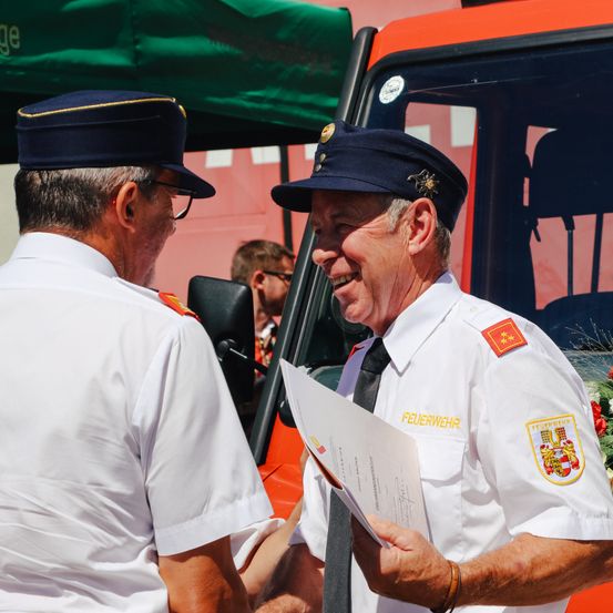 Zwei ältere Männer in weißen Uniformen und Mützen, einer hält ein Dokument, lächeln, während sie neben einem Feuerwehrauto stehen.