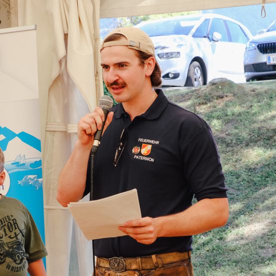 Ein Mann in einem schwarzen Poloshirt hält ein Mikrofon und ein Papier in der Hand, wahrscheinlich spricht er bei einer Veranstaltung. Ein Kind mit einem grünen Shirt schaut zu. Autos sind hinter ihnen geparkt.