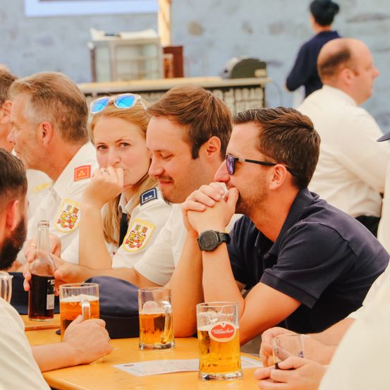 Eine Gruppe von Menschen, darunter zwei Frauen in Uniformen und drei Männer, sitzen an einem Tisch, trinken Bier und unterhalten sich.