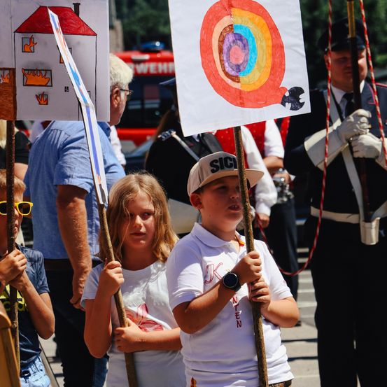 Eine Gruppe von Kindern hält Banner bei einem Umzug, eines mit einem Regenbogen-Ziel und ein anderes mit einem brennenden Haus. Ein Mann und eine Frau im Hintergrund.