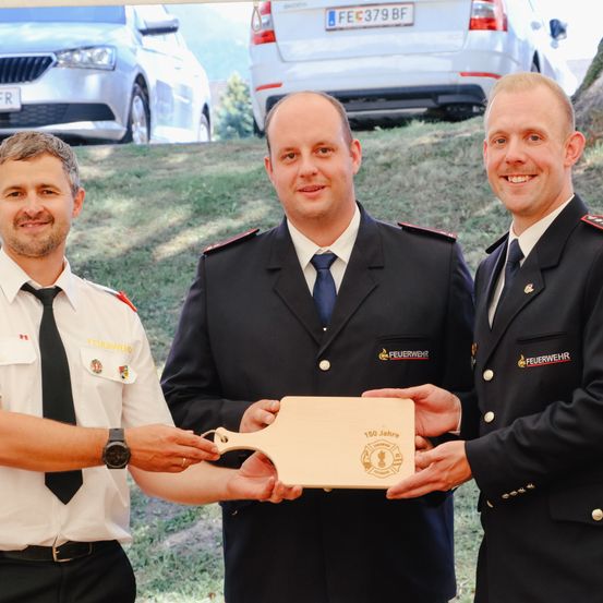 Drei Feuerwehrleute präsentieren eine Plakette mit Logo. Einer hält eine Holzplakette, während die anderen sie an den Kanten halten. Sie stehen auf Gras mit Autos im Hintergrund.